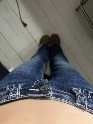 Mörkblå bootcut jeans med slitningar - Säljer ett par mörkblå bootcut jeans med snygga slitningar och kontrastsömmar. Jeansen har klassisk femficksmodell, låg midja och vida ben. Perfekta för dig som gillar en retro och avslappnad stil.