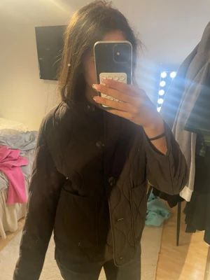 Brun quiltad jacka med knappar - Säljer en brun quiltad jacka med stora knappar framtill och två fickor. Jackan har en rak passform och hög krage, perfekt för lager på lager. Materialet är lätt vadderat och ger en stilren look till vardagsoutfiten.