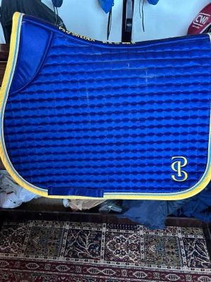 Blått schabrak PS of Sweden - Säljer ett blått schabrak från PS of Sweden med gul kant och broderad text. Schabraket har quiltad struktur och en gul logga i hörnet. Perfekt för dig som vill ha en snygg och färgglad detalj till din hästutrustning.