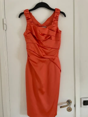 Snygg klänning - Snygg orange kort klänning med breda axelband och draperade detaljer framtill. Klänningen har en figurnära passform och är tillverkad i ett glansigt material!!