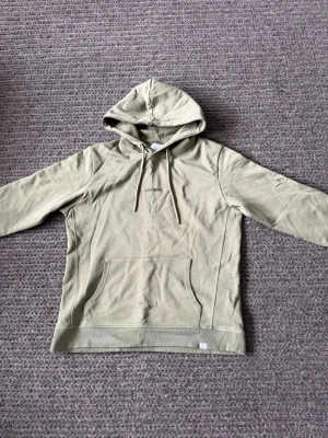 Beige hoodie med magficka och huva - Passar M/L. Snygg beige hoodie med huva och dragsnören. Framtill finns en klassisk magficka och diskret broderad text på bröstet. Tillverkad i mjukt bomullsliknande material, perfekt för en avslappnad stil.