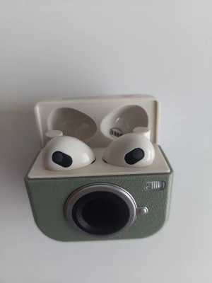 Trådlösa vita in-ear hörlurar med kamerafodral - Säljer ett par trådlösa vita in-ear hörlurar med ett unikt grönt laddningsfodral som ser ut som en retro kamera. Hörlurarna är släta och har svarta detaljer vid högtalaren. Perfekt för dig som vill ha något snyggt och annorlunda till mobilen.