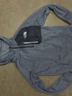 Grå hoodie från The North Face - Snygg grå hoodie från The North Face med svart bröstficka och vit logga. Hoodien har dragkedja vid halsen och en praktisk huva. Perfekt för chill dagar eller när du vill ha en sportig look.