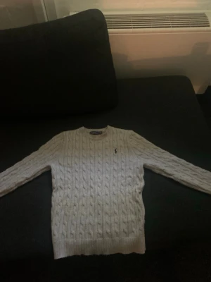 Vit kabelstickad tröja Polo Ralph Lauren - Kabelstickad vit tröja från Polo Ralph Lauren med rund halsringning och diskret broderad logga på bröstet. Perfekt för en klassisk och stilren look.