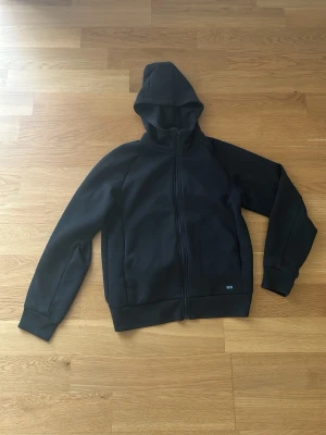 Svart hoodie med dragkedja - En stilren svart hoodie med dragkedja framtill och huva. Tillverkad i ett mjukt och bekvämt material, perfekt för en avslappnad look. Hoodien har ribbade muddar vid ärmar och nederkant samt diskret liten detalj vid nederkanten.