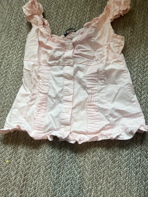 Ljusrosa blus med volangdetaljer - Söt ljusrosa blus med knappar framtill och volangdetaljer vid axelband och nederkant. Blusen har ärmlös design och smockade partier framtill som ger en feminin touch. Perfekt för dig som gillar romantisk stil.