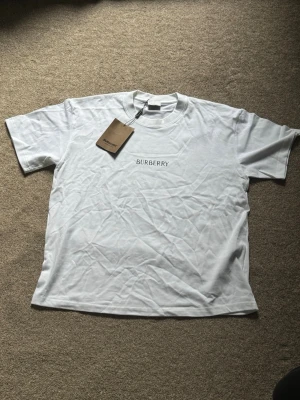 Vit Burberry t-shirt med tryck - Snygg vit t-shirt från Burberry med klassisk logga framtill och stort svart tryck av riddare och Burberry-text på ryggen. T-shirten har rund hals och korta ärmar. Tillverkad i mjuk bomull för en skön känsla. OBS T-shirt är oversized