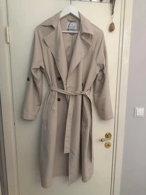 Stradivarius beige trenchcoat med knytskärp - Stilren beige trenchcoat med klassisk krage, dubbelknäppning och knytskärp i midjan. Långa ärmar med knappdetalj och två sidofickor. Perfekt för lager på lager och en tidlös look. Materialet ser ut att vara bomull eller en bomullsblandning.