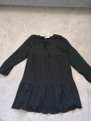 Svart långärmad klänning Samsa - Svart klänning från Samsa Collection i storlek S. Klänningen har lång ärm, volang nedtill och knytning vid halsen. Tillverkad i ett mjukt tyg med lätt struktur. Perfekt för dig som gillar enkel men snygg stil.