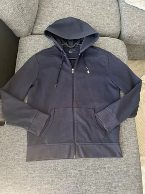 Mörkblå hoodie från Polo Ralph Lauren - Snygg mörkblå hoodie från Polo Ralph Lauren med dragkedja, huva med dragsko och två fickor framtill. Klassisk liten logga broderad på bröstet. Tillverkad i mjukt bomullsmaterial som känns skönt mot huden. Perfekt för en avslappnad och stilren look. Passar s-M