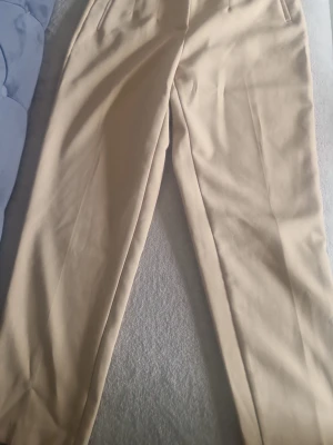 Beige kostymbyxor med hög midja - Snygga beige kostymbyxor med hög midja och raka ben. Byxorna har pressveck framtill och diskreta sidofickor. Perfekta för en clean och stilren look. Materialet ser ut att vara en mjuk och lite tjockare väv, vilket ger en exklusiv känsla.