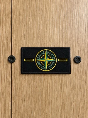 Stone Island tygmärke svart/gul - Säljer ett klassiskt Stone Island tygmärke i svart med broderad gul och grön kompasslogga. Märket har två knapphål på sidorna och passar perfekt att fästa på jackor eller tröjor för en streetwear-look.