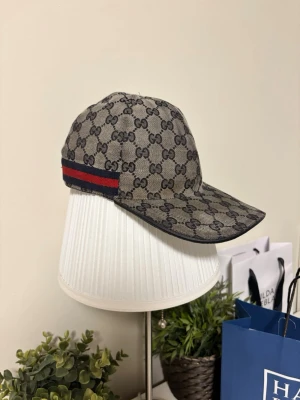 Gucci beige keps med GG-mönster - Säljer en beige keps från Gucci med klassiskt GG-monogram över hela kepsen och en röd-blå rand på sidan. Kepsen har böjd skärm och justerbart spänne bak. Materialet är canvas med insida i tyg. Perfekt för dig som gillar exklusiv streetwear. Kepsen är ÄKTA, kan gå ner lite i pris vid snabb och enkel affär. BARA seriösa bud, Tack på förhand!