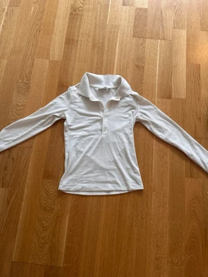 Vit ribbad långärmad polotopp - Säljer en vit ribbad långärmad polotopp med krage och knappar framtill. Toppen har en slim passform och är tillverkad i ett mjukt material som känns skönt mot huden. Perfekt att styla med jeans eller kjol för en clean look.