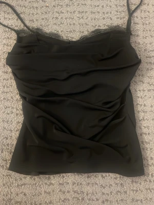 Svart linnetopp med spetskant - Snygg svart linnetopp med tunna axelband och en diskret spetskant upptill. Toppen har en enkel och stilren design som passar perfekt till många olika outfits. Materialet känns mjukt och följsamt.