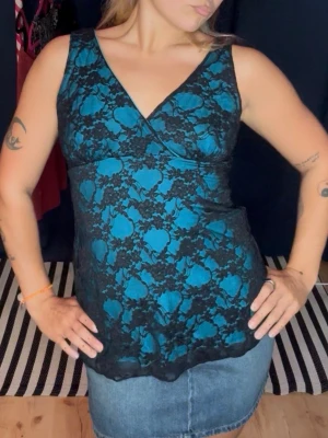 Y2k omlottopp med svart spets - Snygg turkos omlottopp med svart spetsdetalj och blommigt mönster. Toppen är ärmlös och har en v-ringad halsringning som ger en feminin look. Perfekt att styla med jeans eller kjol för en trendig outfit. Storlek L men sitter såhär på mig med storlek S/M. Mycket stretchig ! 