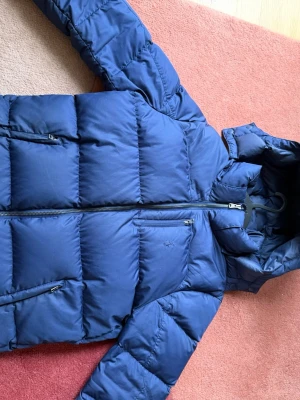 Blå pufferjacka med huva - Säljer en blå pufferjacka med huva och dragkedja framtill. Jackan har två sidofickor med dragkedja och en bröstficka med dragkedja. Den quiltade designen ger en klassisk pufferlook och materialet är syntetiskt för extra värme.