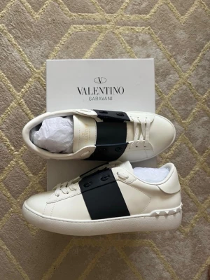 Valentino Garavani open - Snygga vita sneakers från Valentino Garavani med en bred svart remsa över mitten. Skorna har vita snören, rund tå och nitar på hälen. Tillverkade i slätt läder för en clean och exklusiv look. Perfekta för dig som gillar stilrena detaljer. Knappt använda alls