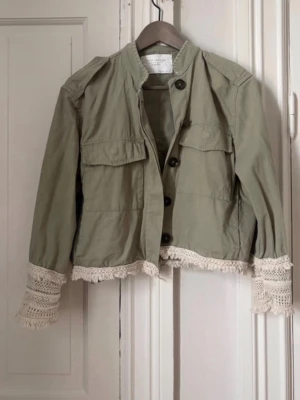 Olivgrön jacka med virkade detaljer - Stilren olivgrön jacka med stora bröstfickor och svarta knappar framtill. Jackan har unika virkade spetsdetaljer i beige vid ärmslut och nederkant som ger en bohemisk touch. Tillverkad i ett lätt bomullstyg, perfekt för lager på lager och trendiga outfits.