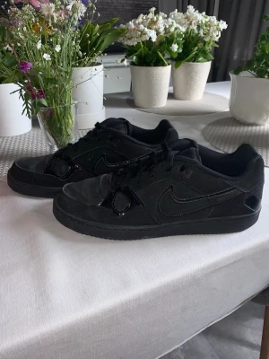 Svarta Nike sneakers med mockadetaljer - Svarta sneakers från Nike med lågt skaft, klassisk swoosh-logga på sidan och svarta skosnören. Skorna har en mix av mocka och syntetmaterial samt diskreta glansiga detaljer vid snörningen. Perfekta för en stilren  look.