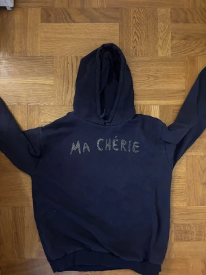 Mörkblå hoodie Ma Chérie Gina Tricot - Mörkblå hoodie från Gina Tricot med texten 'Ma Chérie' i glittriga stenar på bröstet. Klassisk huva och ribbade muddar vid ärmslut och nederkant. Tillverkad i mjukt bomullsmaterial för en skön känsla.