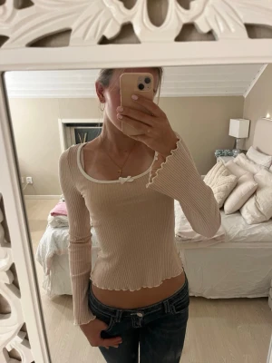 Ribbad topp med rosett - Säljer en söt rosa ribbad topp med lång ärm och vit kant runt den djupa ringningen. Toppen har en liten rosett framtill och vågig kant nertill och vid ärmsluten💗