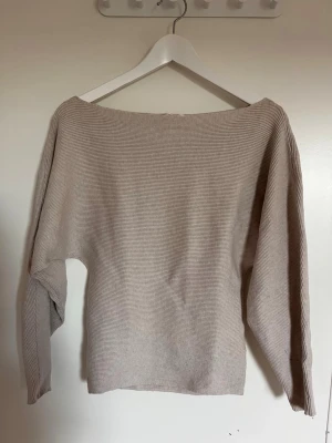 Beige ribbstickad tröja - En stilren beige ribbstickad tröja med bred båturringning och långa ärmar. Tröjan har en avslappnad passform och är tillverkad i ett mjukt, tunt stickat material som passar perfekt till lager på lager. Enkel att matcha med olika outfits.