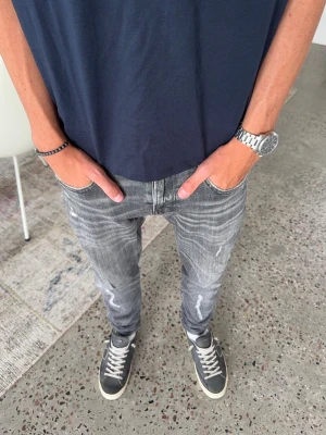 Grå slitna slim fit jeans från replay - Säljer ett par grå slim-fit jeans med slitningar och tvättade detaljer. Jeansen har fem fickor, dragkedjegylf och smal passform som sitter skönt längs benen. Perfekta för en avslappnad och trendig look. W30 L30 men passar om man har L32 också