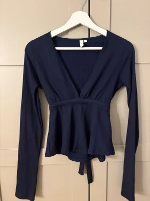 Mörkblå omlottopp med knytband - Snygg mörkblå omlottopp med långa ärmar och v-ringning. Toppen har ett knytband i midjan som ger en fin siluett och är tillverkad i ett ribbat material. Perfekt att styla med jeans eller kjol för en trendig look.