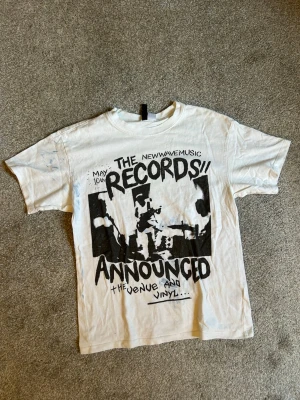 Vit t-shirt med tryck  - Säljer en vit t-shirt med svart tryck framtill i texten 'The Records Announced' samt blå målarfläckar som är en del av tröjan. T-shirten har rund hals och korta ärmar. 