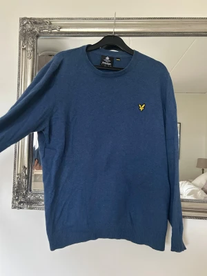 Blå stickad tröja från Lyle & Scott - Snygg blå stickad tröja från Lyle & Scott med rund halsringning och den klassiska gula loggan broderad på bröstet. Tröjan har ribbade muddar vid ärmslut och nederkant. Perfekt till lager på lager och enkel att matcha.