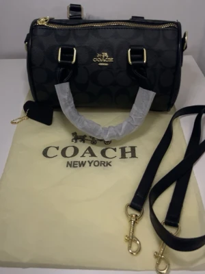Svart rowan handväska från Coach - Snygg svart handväska från Coach med klassiskt monogrammönster och guldfärgade detaljer. Väskan har både handtag och avtagbar axelrem, samt dragkedja upptill. Tillverkad i tåligt konstläder och levereras med dustbag.
