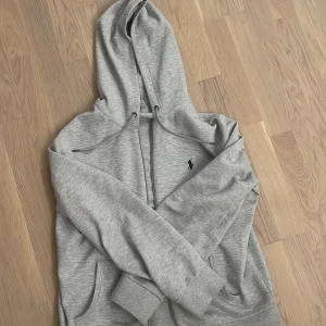 Grå zip hoodie från Ralph Lauren - Säljer en stilren grå hoodie från Ralph Lauren med dragkedja och huva. Den har en liten broderad logga på bröstet och är tillverkad i ett mjukt material. Har bara använt tröjan ett par få gånger