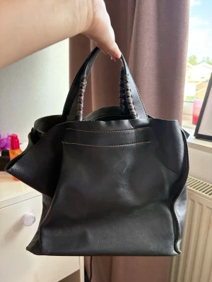 Svart läderhandväska med flätade handtag - Snygg svart handväska i mjukt läder med flätade handtag och kontrastsömmar. Väskan har ett rymligt huvudfack och en innerficka med dragkedja. Perfekt för dig som vill ha en stilren och praktisk väska till vardagen.