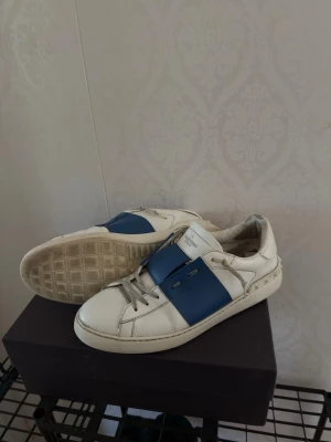 Valentino Garavani blåa sneakers  - Säljer ett par riktigt snygga Valentino Garavani sneakers i vitt med blåa detaljer.  En riktigt clean och trendig sneaker som passar till både jeans, shorts och lite mer uppklädda outfits. Den vita/blå färgkombinationen gör dem dessutom väldigt lätta att matcha.  Skorna är använda och har vissa tecken på användning, se bilder för skick. Hör gärna av dig om du vill ha fler bilder eller har någon fundering.