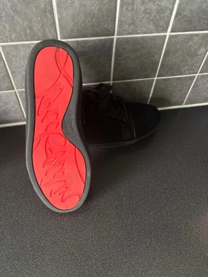 Loubs  sneakers med röd sula - Snygga svarta sneakers med klassisk låg profil och en riktigt cool röd sula. Skorna har svarta skosnören och ovandel i mockaliknande material. Perfekta för dig som vill sticka ut med en unik detalj under foten.