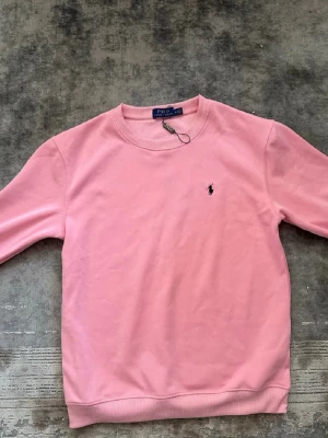 Rosa sweatshirt från Polo Ralph Lauren - Snygg rosa sweatshirt från Polo Ralph Lauren med klassisk rund halsringning och broderad logga på bröstet. Tröjan har ribbade muddar vid ärmslut och nederkant. Perfekt för en avslappnad och trendig look.