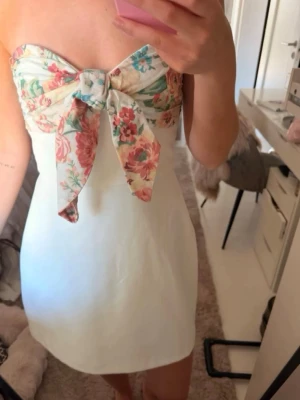 Vit kort klänning med blommig rosett från Zara - Supersöt vit kort klänning med axelbandslös design och stor blommig rosett framtill. Överdelen har ett färgglatt blommönster i rosa, grönt och blått. Perfekt för sommardagar och fester. Klänningen har en feminin och trendig look. Klänningen är slutsåld och näst intill oanvänd i storlek M
