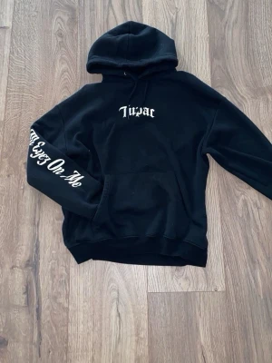 Svart 2Pac hoodie (L) - Säljer en svart hoodie från Pull&Bear med 2Pac-tryck på bröstet och texten 'All Eyes On Me' på ena ärmen. Klassisk känguruficka framtill och huva med dragsko. Perfekt för dig som gillar streetwear och vill ha en unik hoodie med hiphop-vibe.