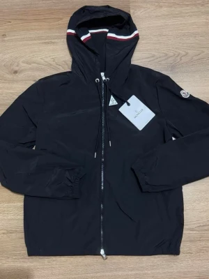 Svart vindjacka med huva från Moncler - Snygg svart vindjacka från Moncler med hel dragkedja, huva med röd och vit rand, samt dragsko. Jackan har Moncler-logga på ärmen och elastiska muddar vid ärmslut och nederkant. Perfekt för blåsiga dagar och enkel att matcha. Kommer utan tagg