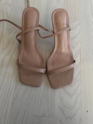 Beige sandaletter med fyrkantig tå - Snygga sandaletter i beige med smala remmar och fyrkantig tå. Skorna har en minimalistisk design och låg klack, perfekta för en trendig look. Materialet ser ut att vara konstläder.