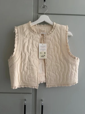 Beige väst från Sézane - Supersöt beige quiltad väst från Sézane med volangkant runt halsen och ärmhålen. Väldigt boho chic! 
