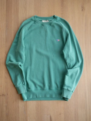 Grön sweatshirt från Maison Kitsuné - Snygg grön sweatshirt från Maison Kitsuné med broderad rävlogga i blått, vitt och rött på bröstet. Tröjan har rund halsringning, raglanärmar och ribbade muddar vid ärmslut och nederkant. Perfekt för en avslappnad och trendig stil.