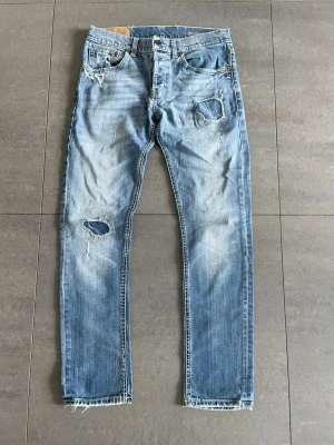 Dondup jeans - Säljer ett par ljusblå jeans med skinny fit och slitna detaljer, inklusive hål på knät och låret. Jeansen har råa kanter vid bensluten och klassisk femficksdesign. Perfekta för en avslappnad och trendig look.