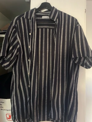 Randig skjorta från Jack & Jones - Snygg kortärmad skjorta från Jack & Jones med svart bas och vita vertikala ränder. Skjortan har klassisk krage, knäppning framtill och luftig passform. Perfekt till sommaren och enkel att styla med jeans eller shorts.