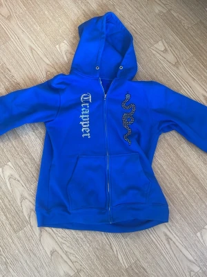 Blå Mads by Trapper hoodie - Säljer en blå hoodie med dragkedja, stor huva och fickor framtill. På bröstet finns ett tryck med texten 'Trapstar' och en orm i glittrande stenar. Perfekt för dig som gillar streetwear och vill sticka ut.