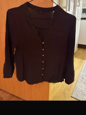 Brown Button-Up Blouse - This is a women's brown blouse with long sleeves and a v-neckline. It features a row of gold colored buttons down the front. The fabric appears to be lightweight and flowy, making it suitable for both casual and semi-formal occasions.