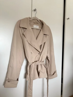 Kort beige trenchcoat med knytskärp - Stilren kort trenchcoat i beige med bred krage, dubbelknäppning och knytskärp i midjan. Jackan har dekorativa knappar vid ärmsluten och klassiska detaljer som axelklaffar saknas. Perfekt för dig som gillar en enkel och tidlös look.