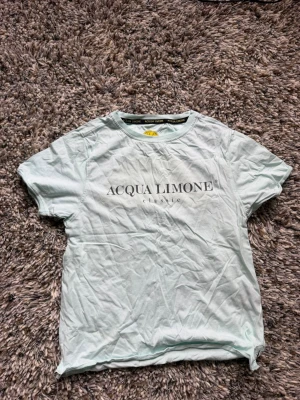 Ljusblå Acqua Limone t-shirt - Säljer en ljusblå t-shirt från Acqua Limone med tryck fram och bak. Framsidan har logotypen och baksidan har en stor gul citron-grafik. Klassisk passform och korta ärmar, perfekt till sommaren.