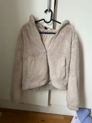 Beige teddyjacka med huva - Supermjuk och fluffig teddyjacka i beige färg med dragkedja framtill och två fickor. Jackan har en mysig huva och är fodrad på insidan. Perfekt för kyliga dagar när du vill vara både varm och stylish. Från H&M
